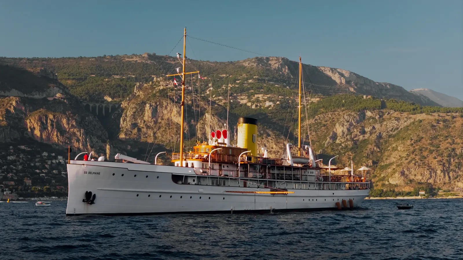 SS Delphine at anchor in golden light below a steep wooded headland.