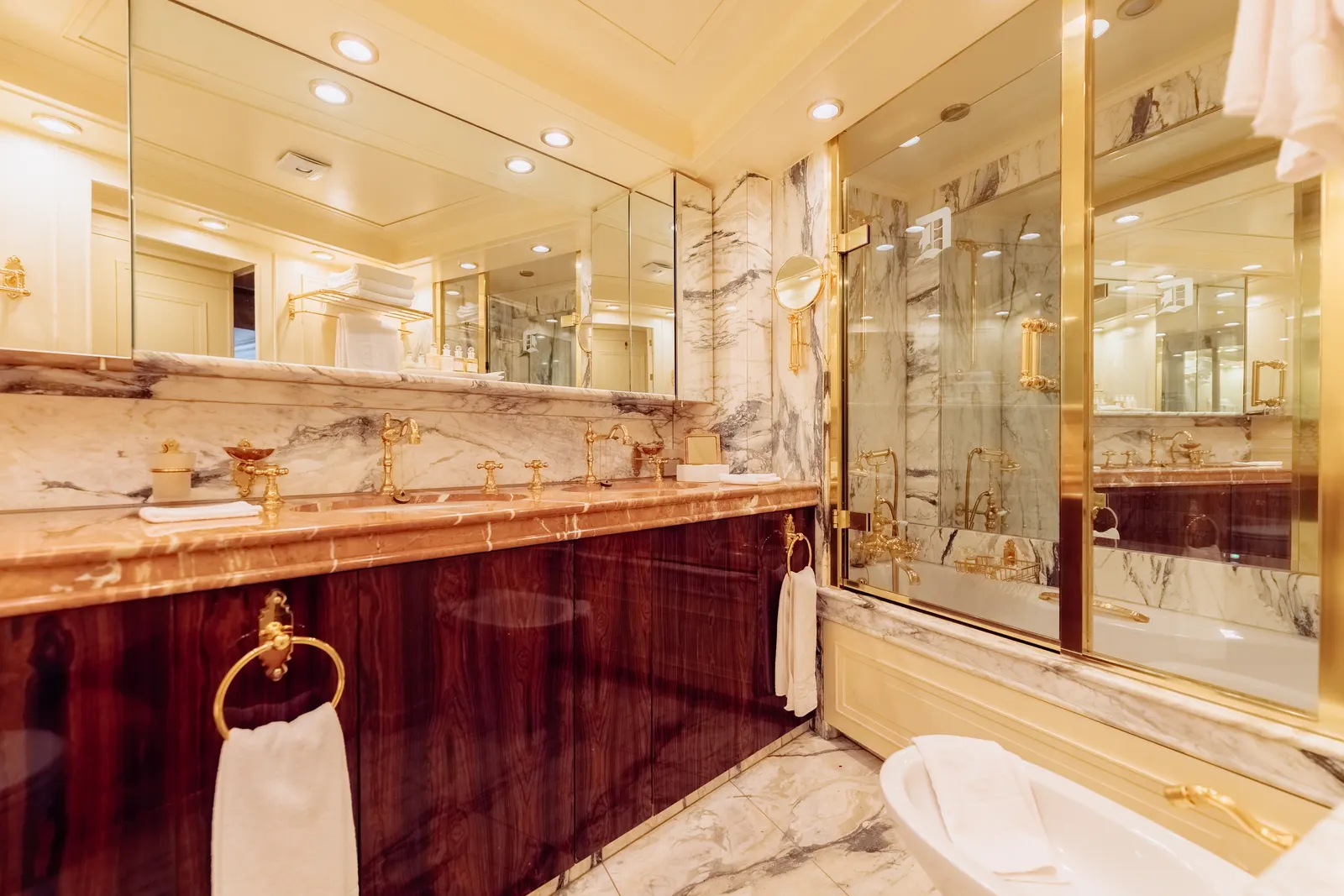 A bath set into marble beside a mirrored wall and a mahogany cabinet.