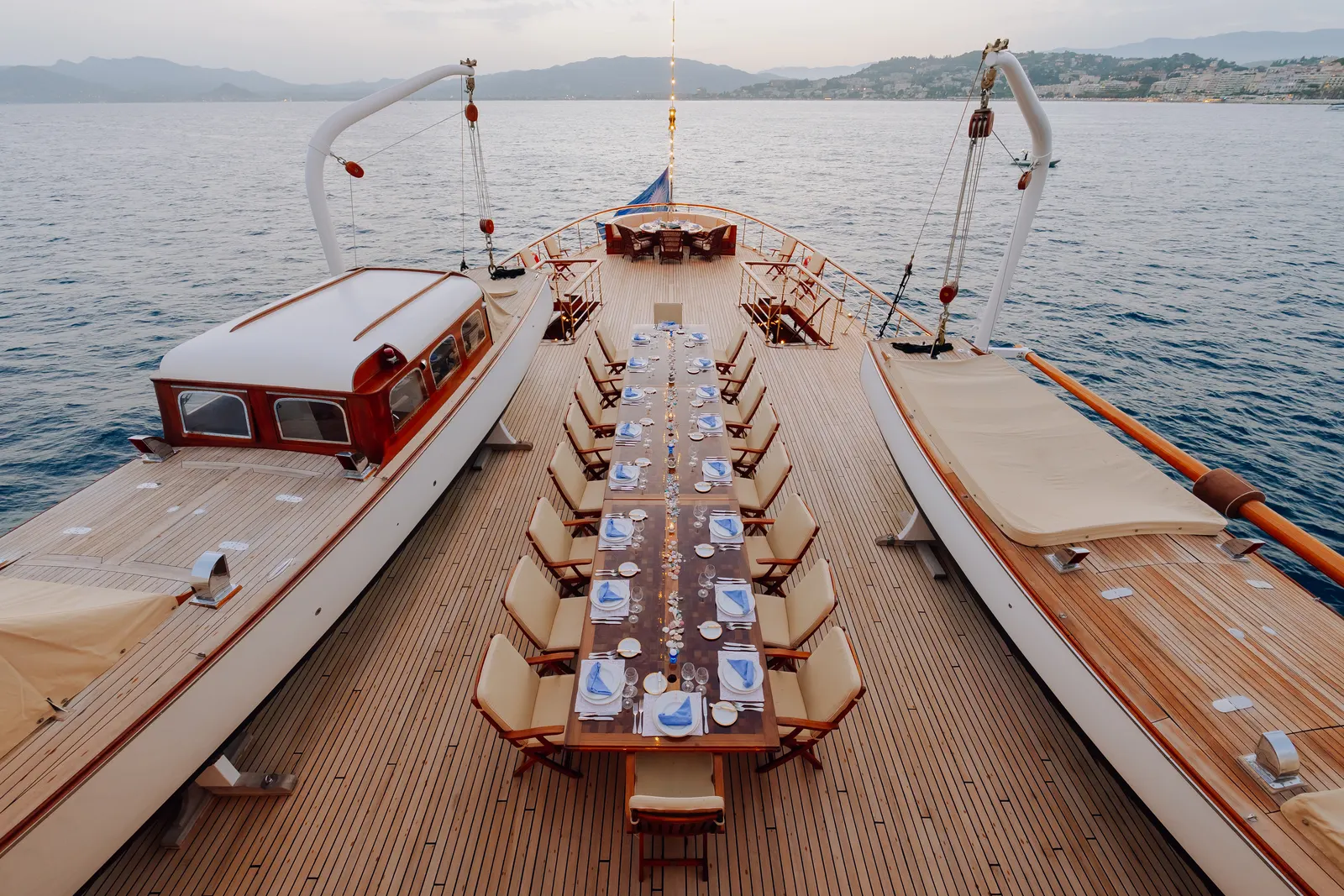 A long table laid for dinner down the teak deck of SS Delphine, seen from above with the tender cradled alongside.