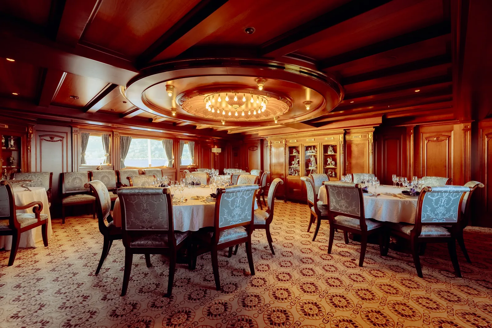 The dining room of SS Delphine laid for service beneath its chandelier, tables set along panelled walls.