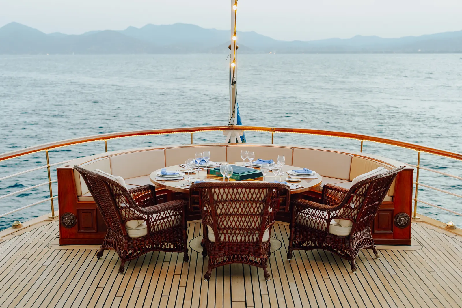 A round table set for four in wicker chairs on the foredeck, open sea beyond the rail.