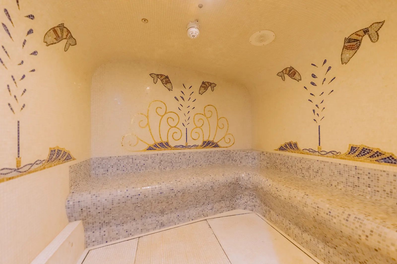 A mosaic lined steam room with a curved bench and painted fish above it.