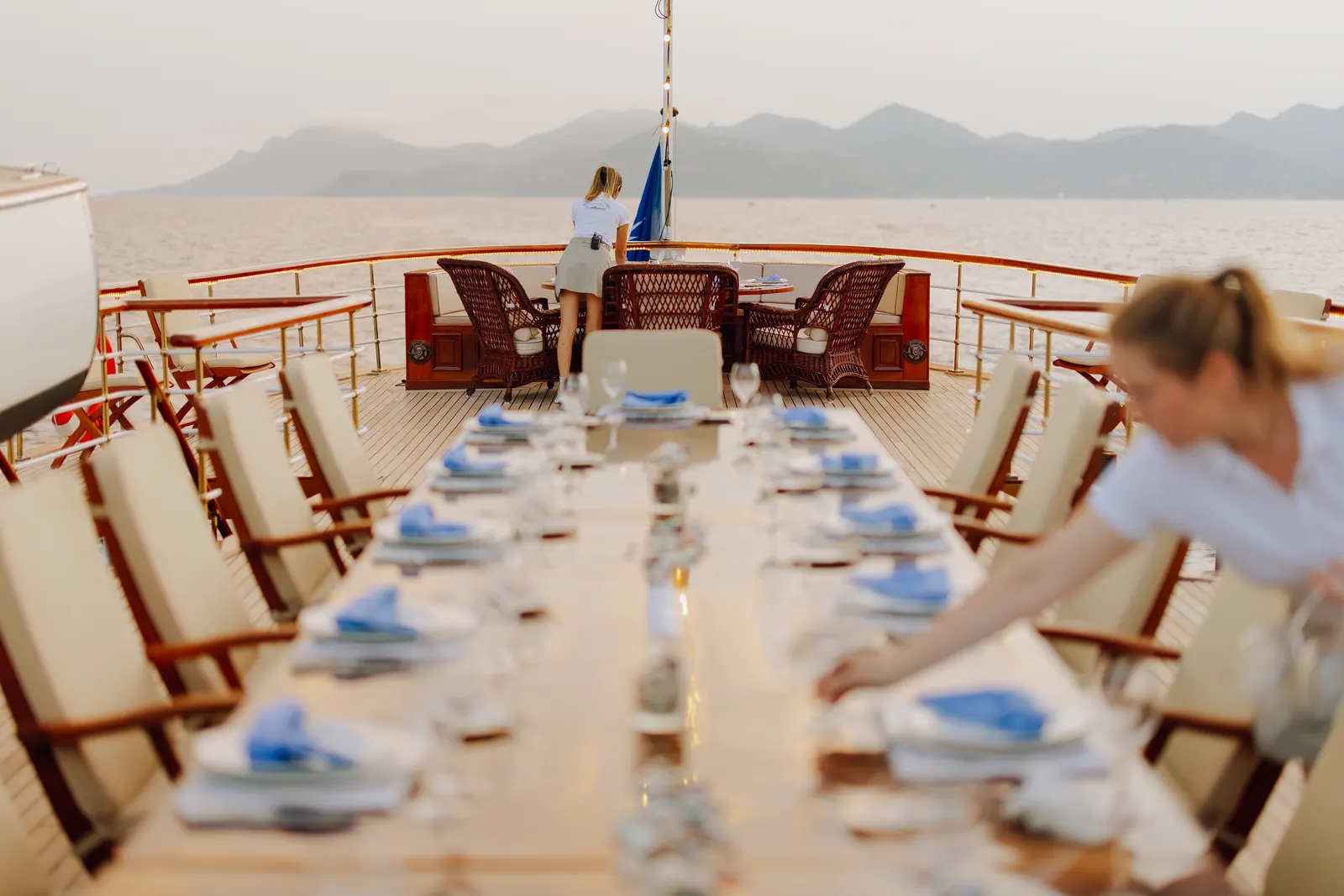 A stewardess laying the long deck table, mountains across the water behind her.