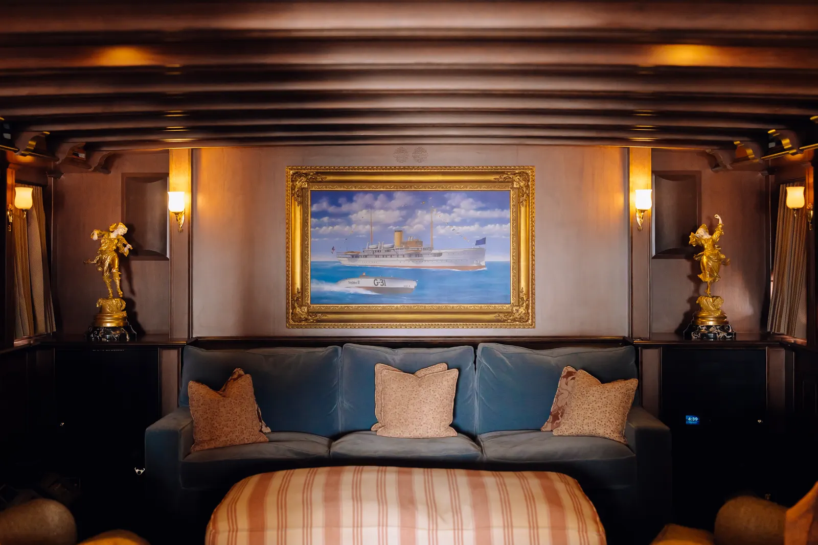 A small lounge aboard SS Delphine with a blue sofa beneath a gilt framed painting of the yacht.