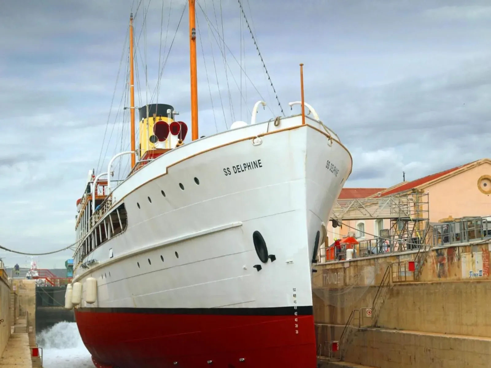 SS Delphine sitting dry in the dock during her refit, her red hull and white superstructure seen from the quayside.